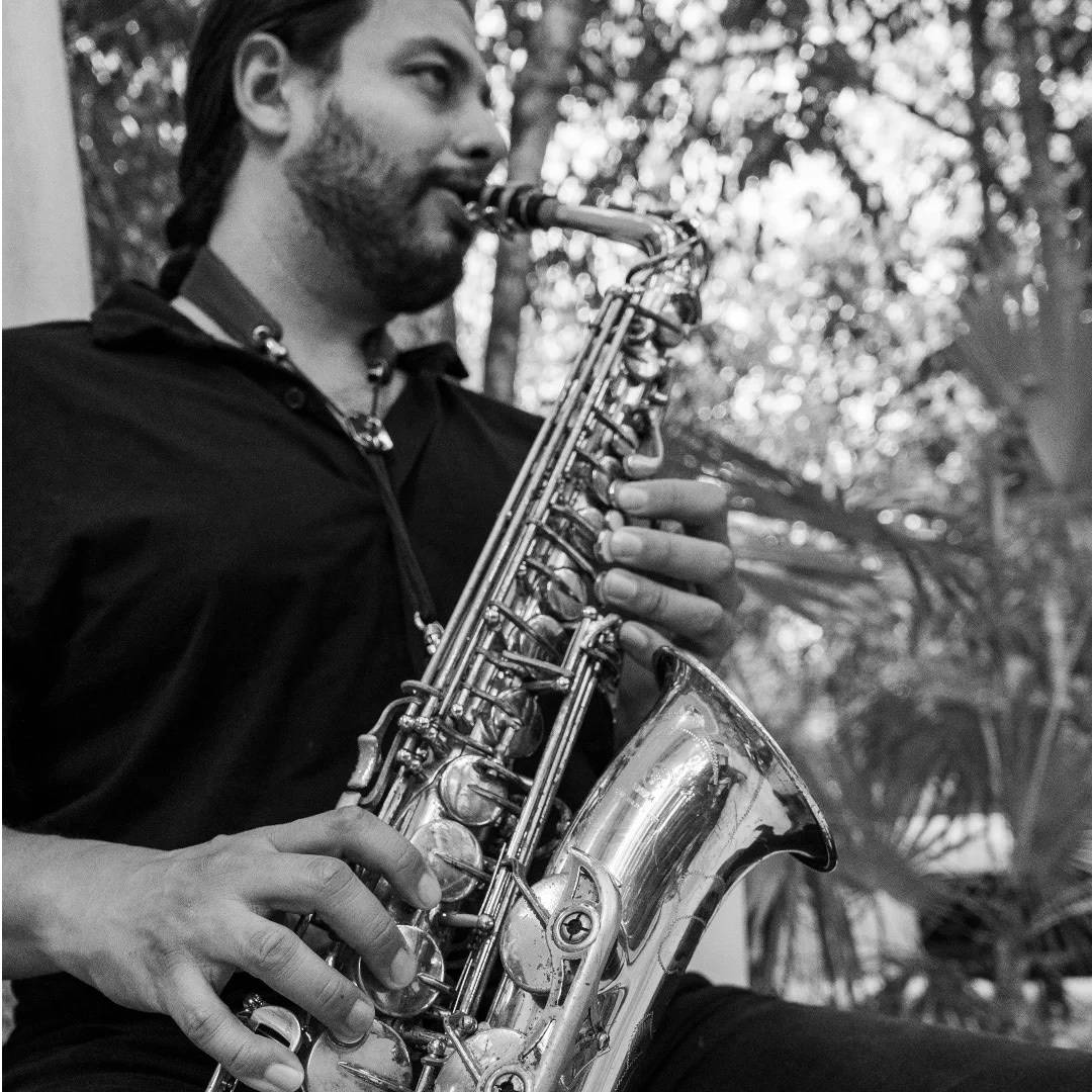 Agustín Yañez playing the alto saxophone, in black and white, with greenery behind him.