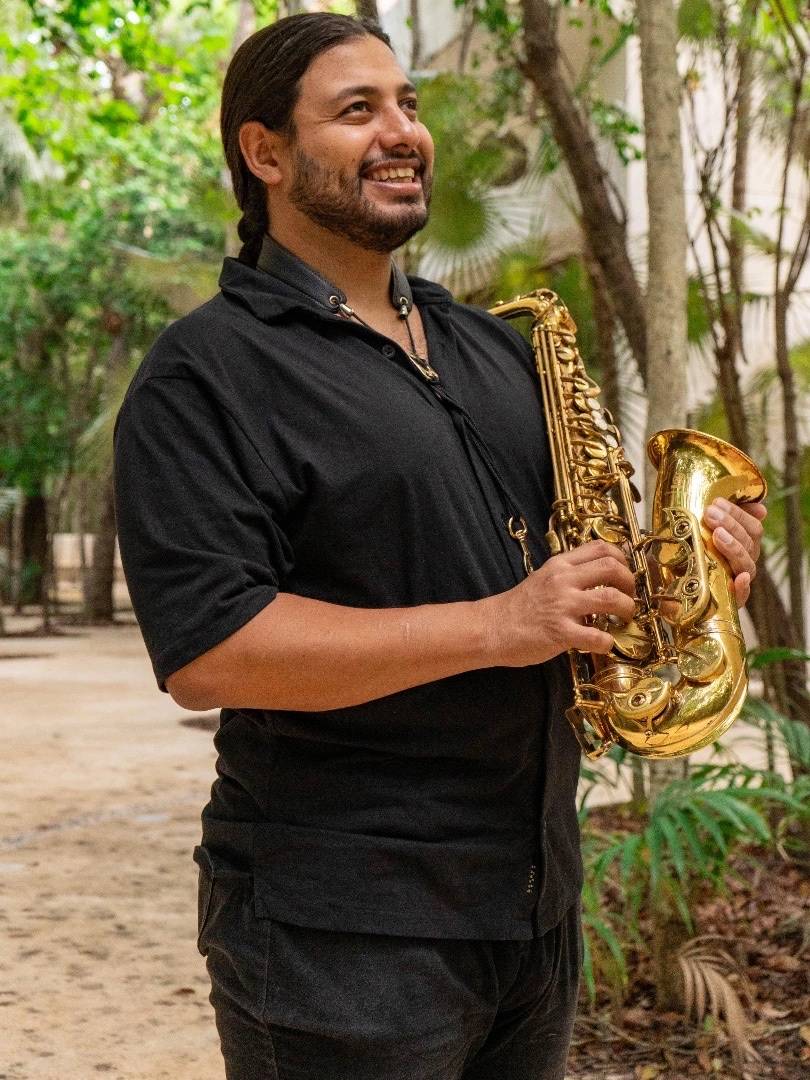 Agustín Yañez smiling, standing with his alto saxophone among the greenery of Cozumel.